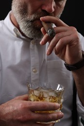 Bearded man with glass of whiskey smoking cigar against black background, closeup