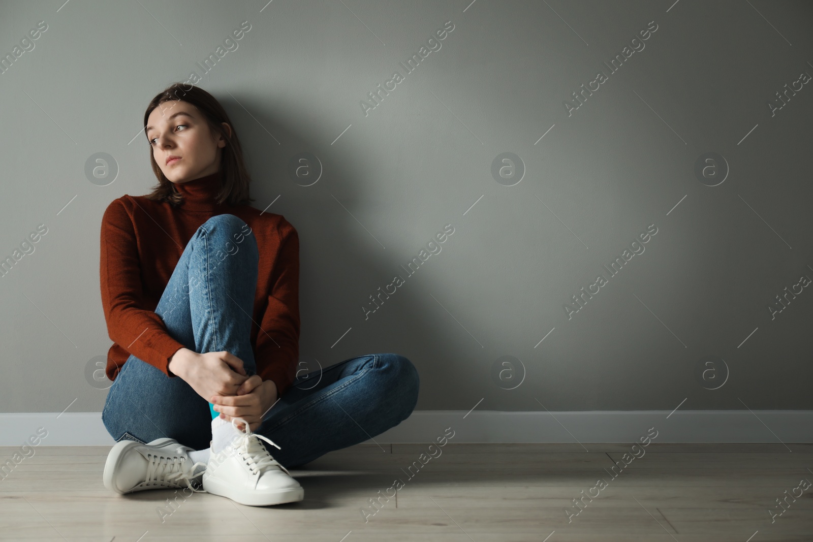 Photo of Sad young woman sitting on floor near grey wall indoors, space for text