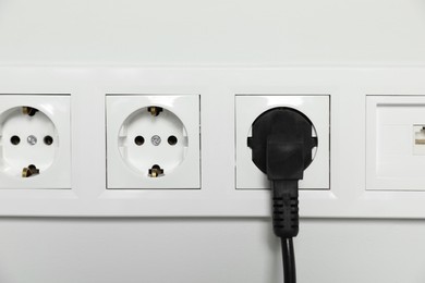 Power sockets with inserted plug on white wall, closeup. Electrical supply