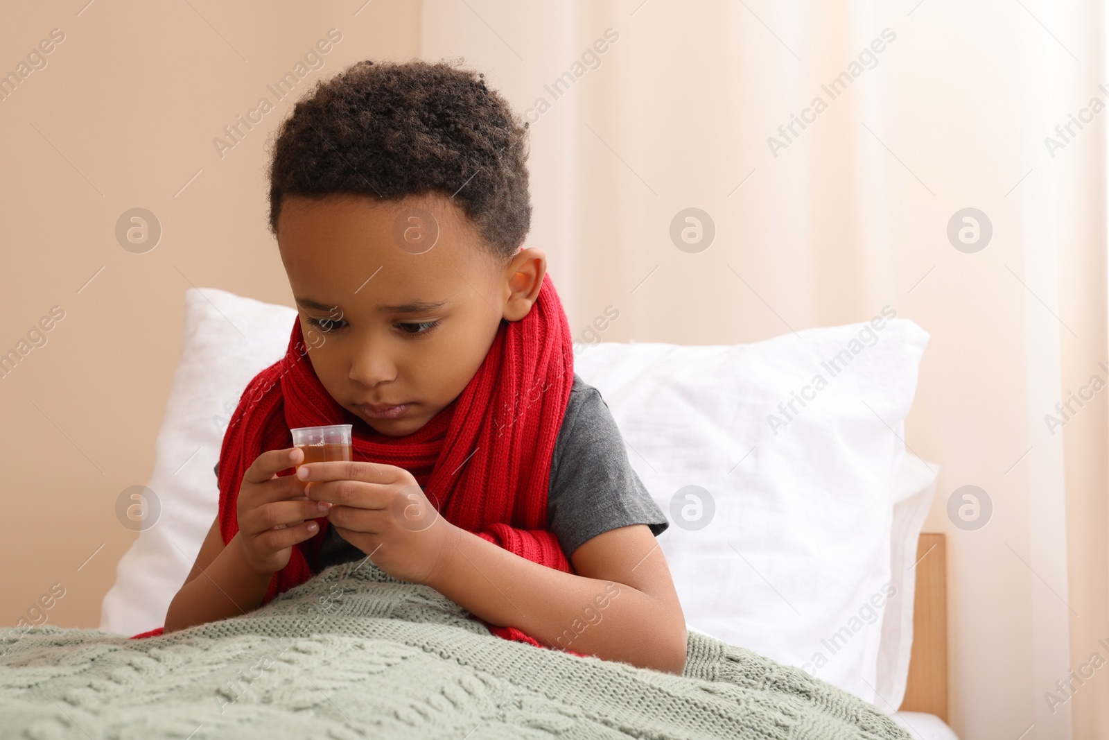 Photo of African-American boy taking cough syrup on bed at home, space for text. Cold medicine