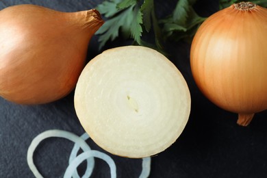 Whole and cut onions with parsley on black textured table, flat lay