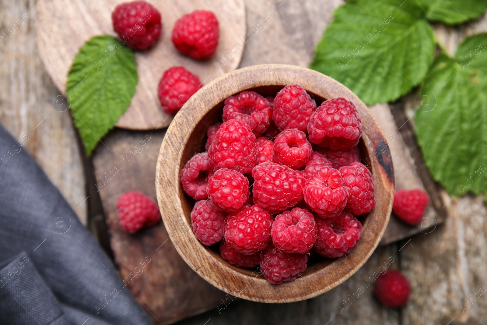 Photo of Bowl with fresh ripe raspberries and green leaves on wooden table, flat lay