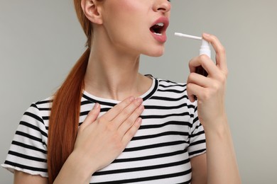 Young woman using throat spray on grey background, closeup