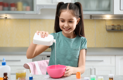 Photo of Cute little girl pouring glue into bowl at table in kitchen. DIY slime toy