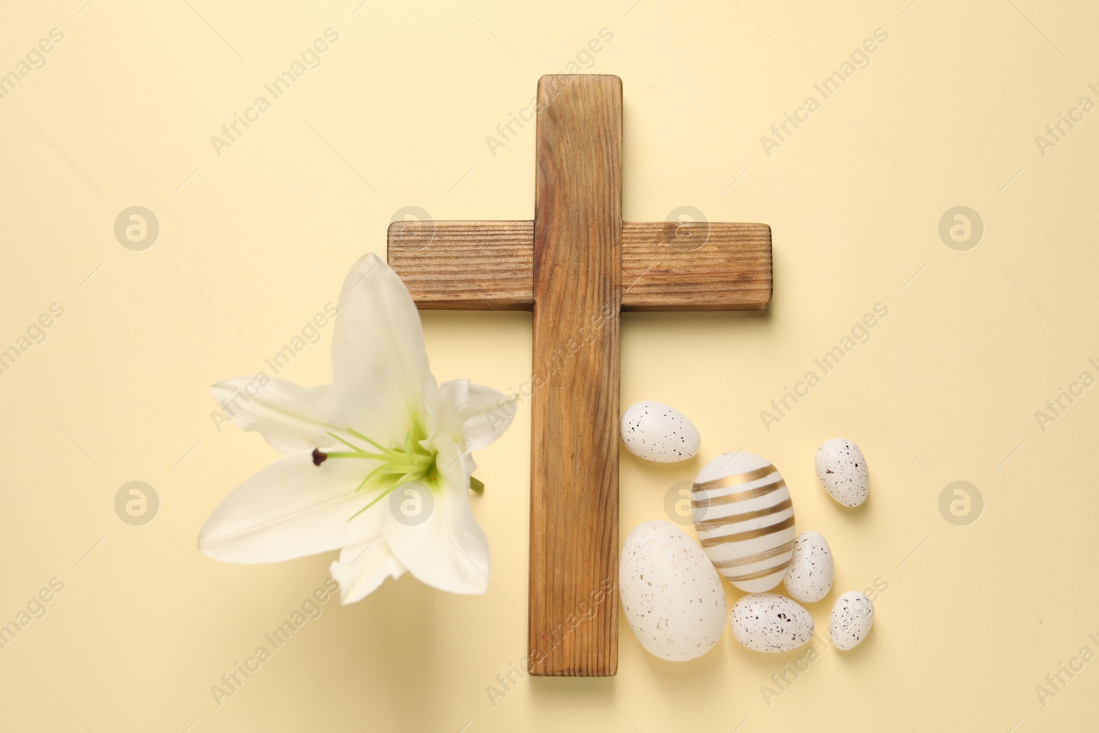 Photo of Wooden cross, painted Easter eggs and lily flower on pale yellow background, flat lay
