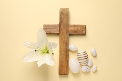 Wooden cross, painted Easter eggs and lily flower on pale yellow background, flat lay