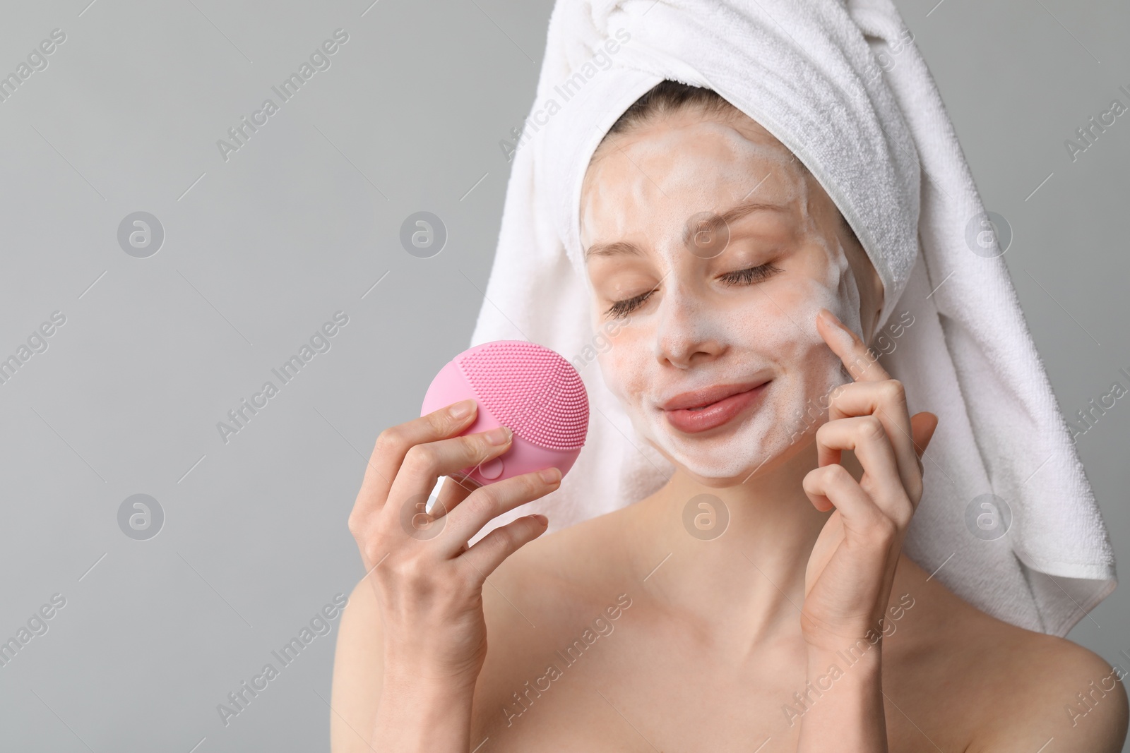 Photo of Young woman washing face with brush and cleansing foam on grey background. Space for text