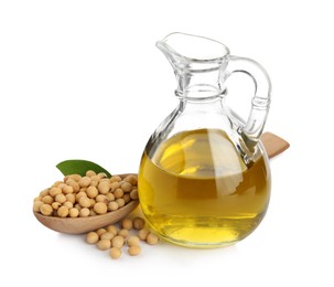 Photo of Glass jug of oil, wooden spoon and soybeans on white background