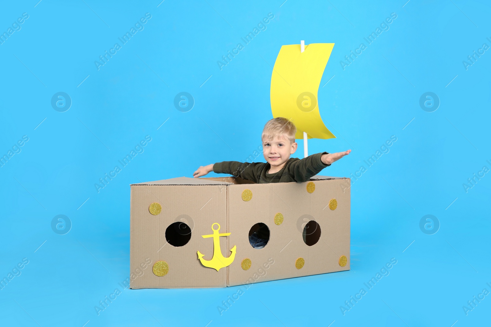 Photo of Little child playing with ship made of cardboard box on light blue background