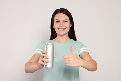 Beautiful young woman holding tin can with beverage and showing thumb up on light grey background