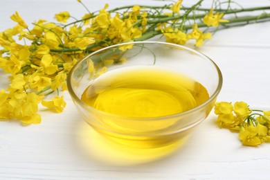 Rapeseed oil in bowl and beautiful yellow flowers on white wooden table, closeup