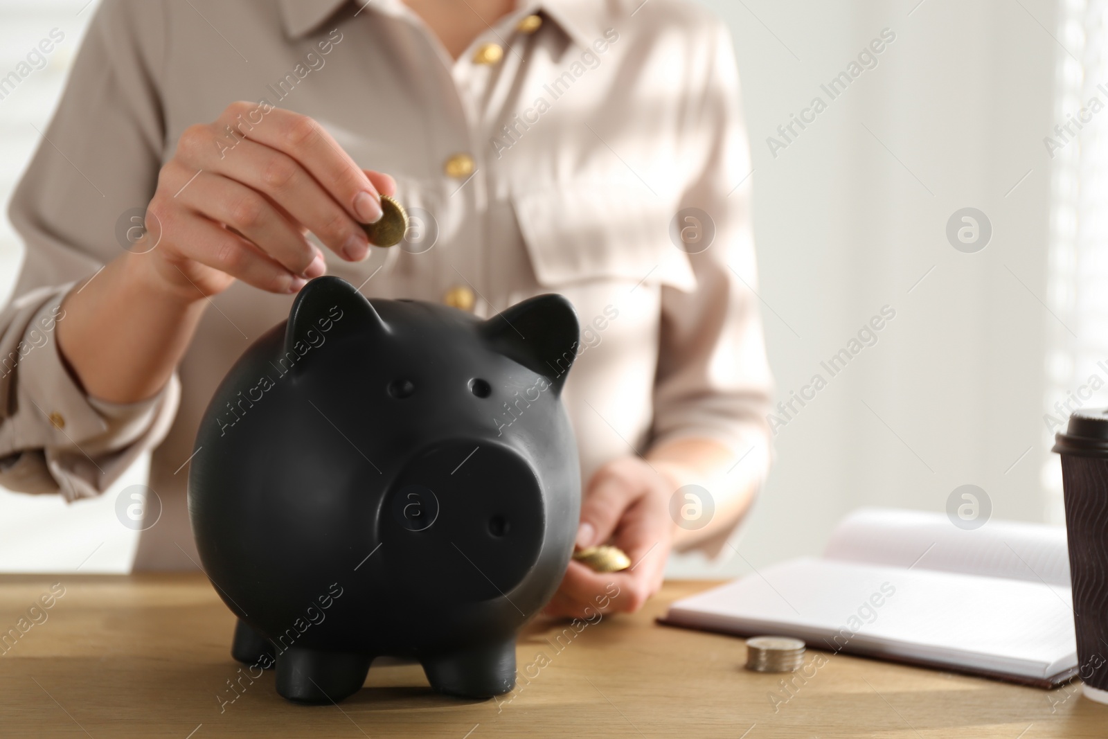 Photo of Woman putting money into piggy bank at wooden table indoors, closeup