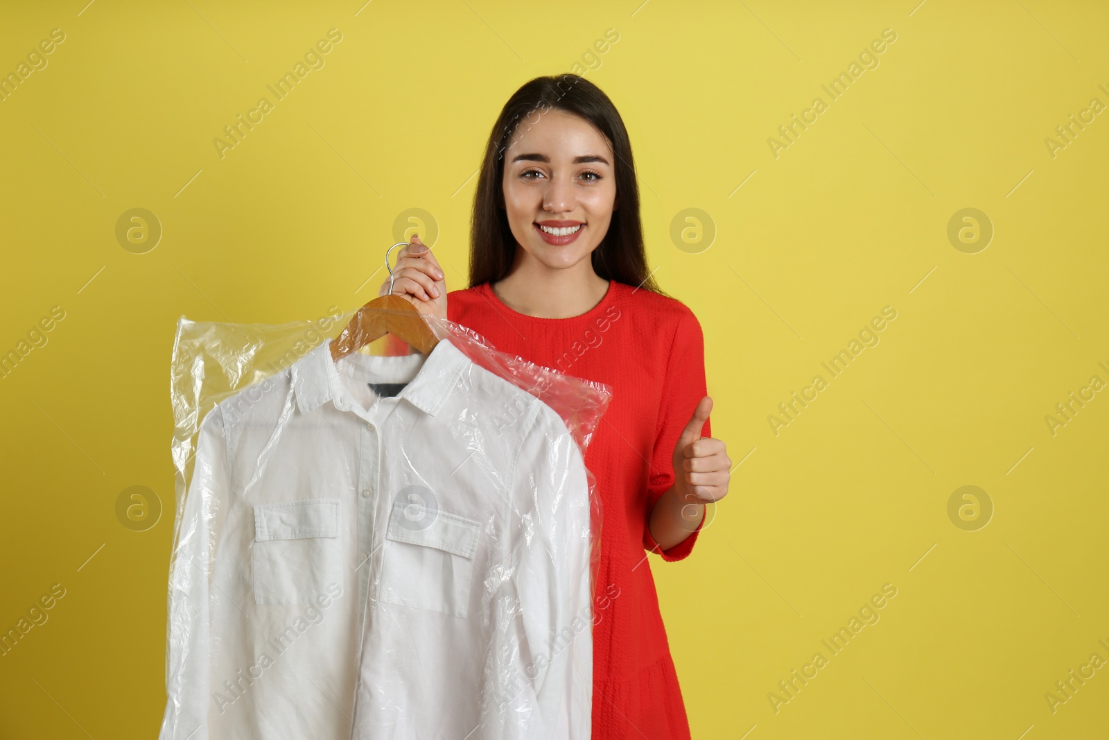Photo of Young woman holding hanger with shirt in plastic bag on yellow background. Dry-cleaning service