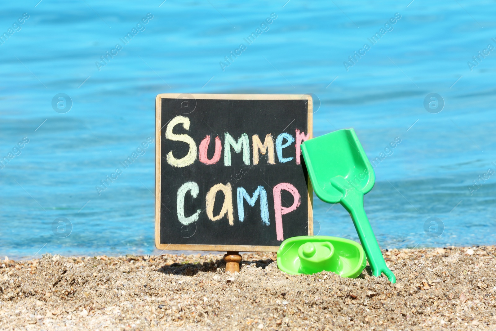 Photo of Little blackboard with text SUMMER CAMP and toys on sand near sea