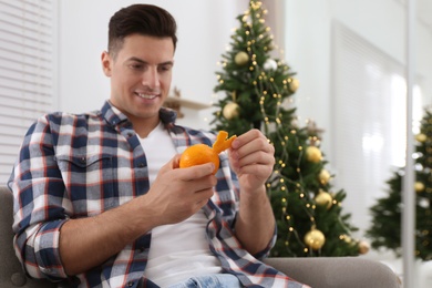 Happy man peeling tangerine near Christmas tree indoors