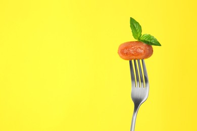 Fork with tasty fried sausage and basil leaves on yellow background. Space for text