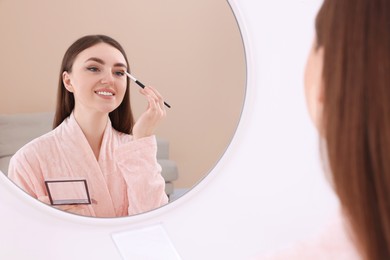 Photo of Beautiful young woman applying eyeshadow with brush near mirror indoors