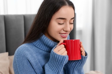 Photo of Happy young woman with red ceramic mug on bed at home