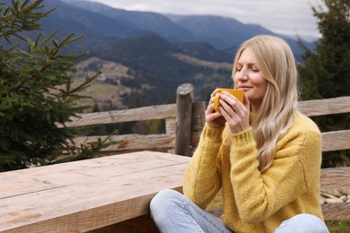 Photo of Young woman with mug of hot drink at wooden table in mountains