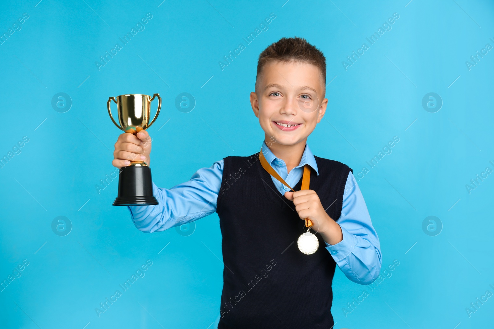 Photo of Happy boy in school uniform with golden winning cup and medal on blue background