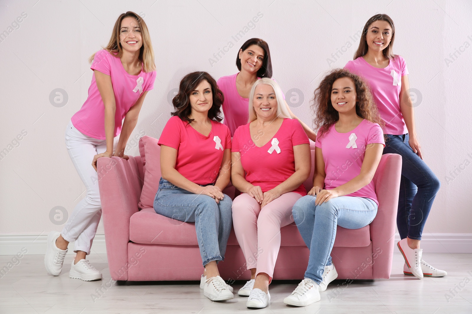 Photo of Group of women with silk ribbons sitting on sofa against light wall. Breast cancer awareness concept