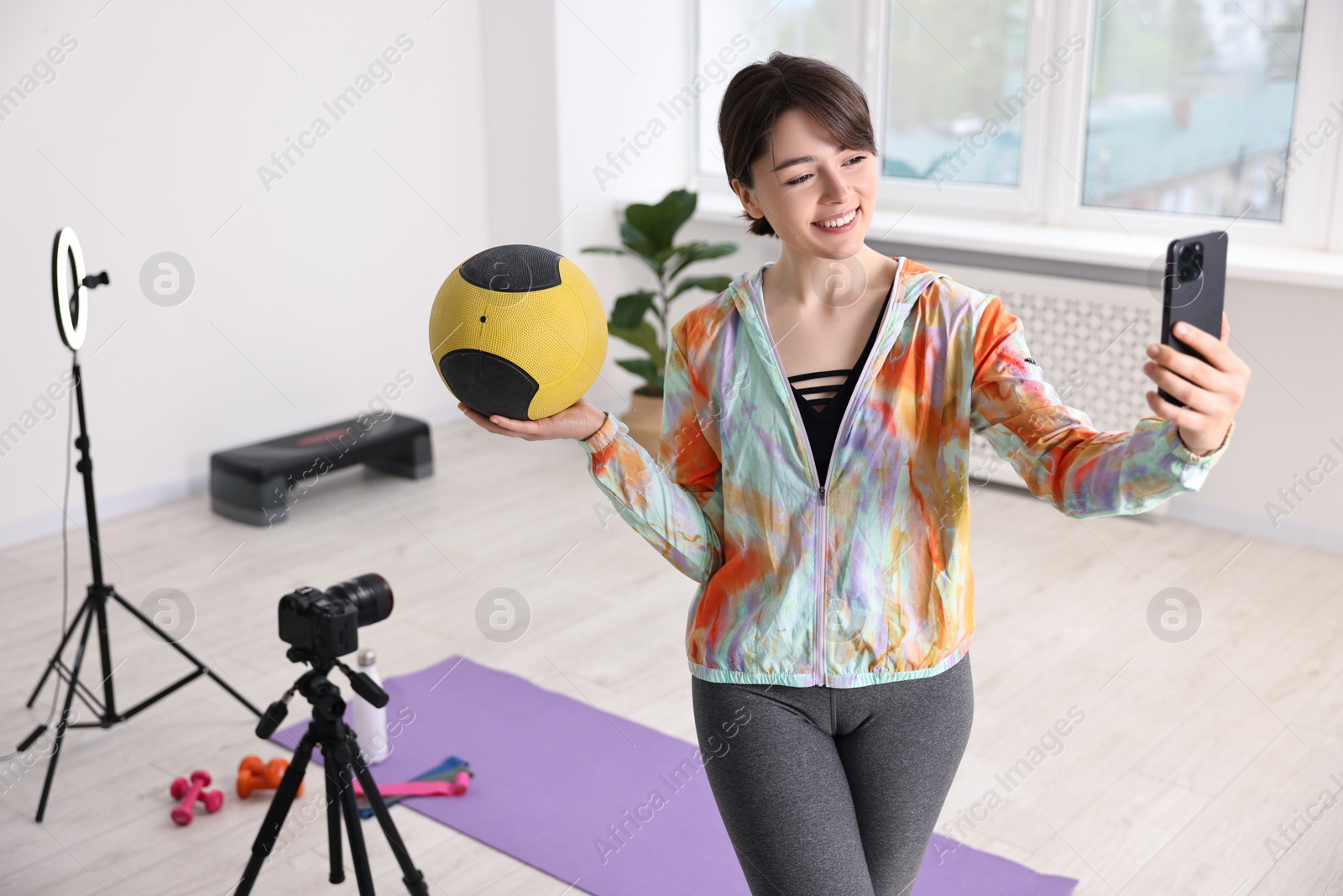 Photo of Happy sports blogger holding medicine ball while streaming online fitness lesson with smartphone at home