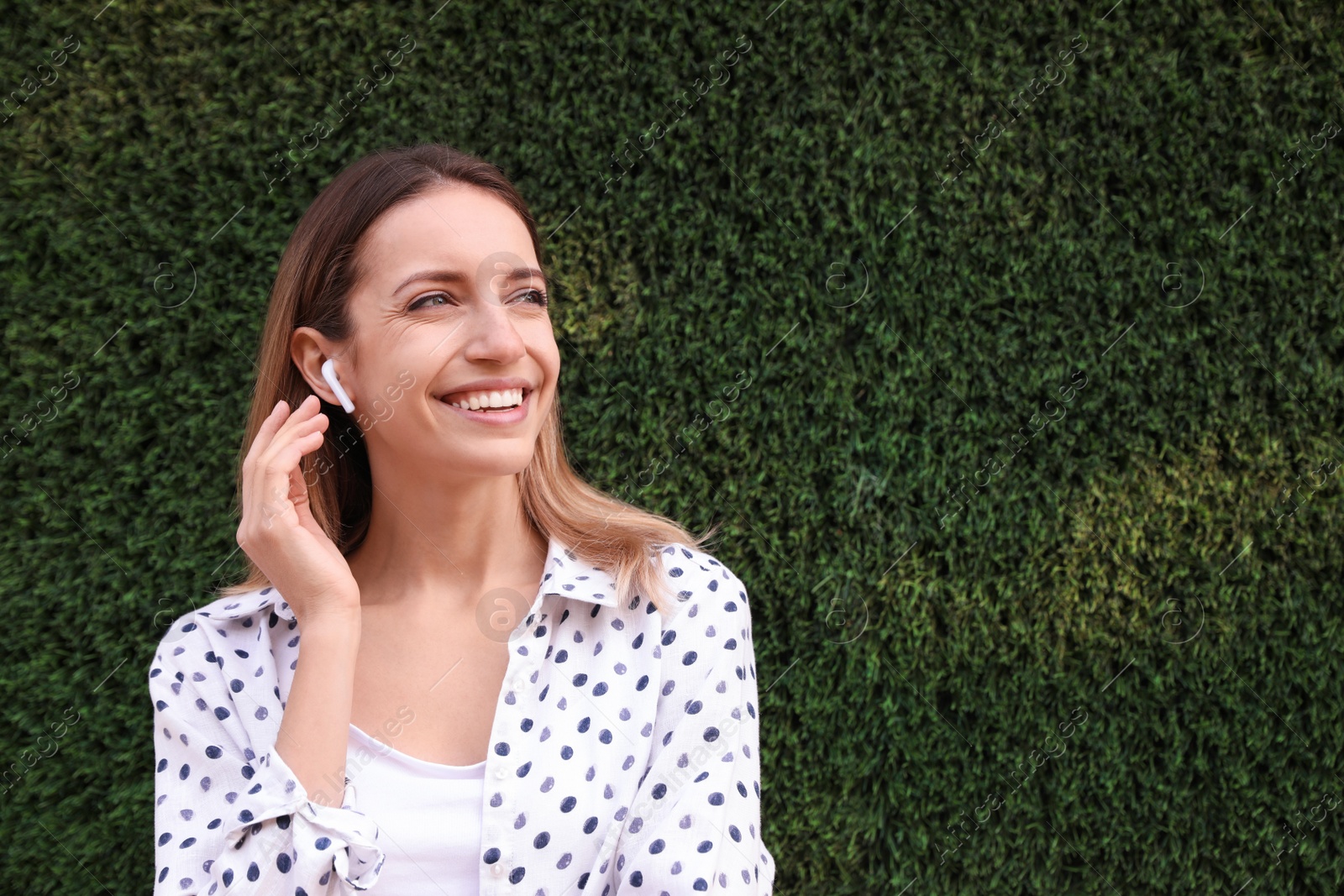 Photo of Young woman with wireless headphones listening to music near green grass wall. Space for text