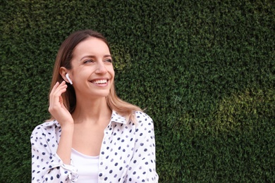 Young woman with wireless headphones listening to music near green grass wall. Space for text