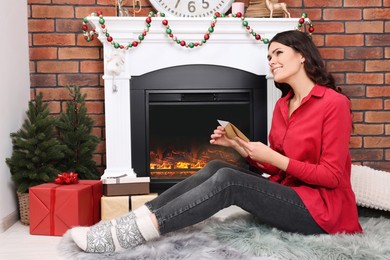 Photo of Young woman with greeting card sitting near fireplace indoors