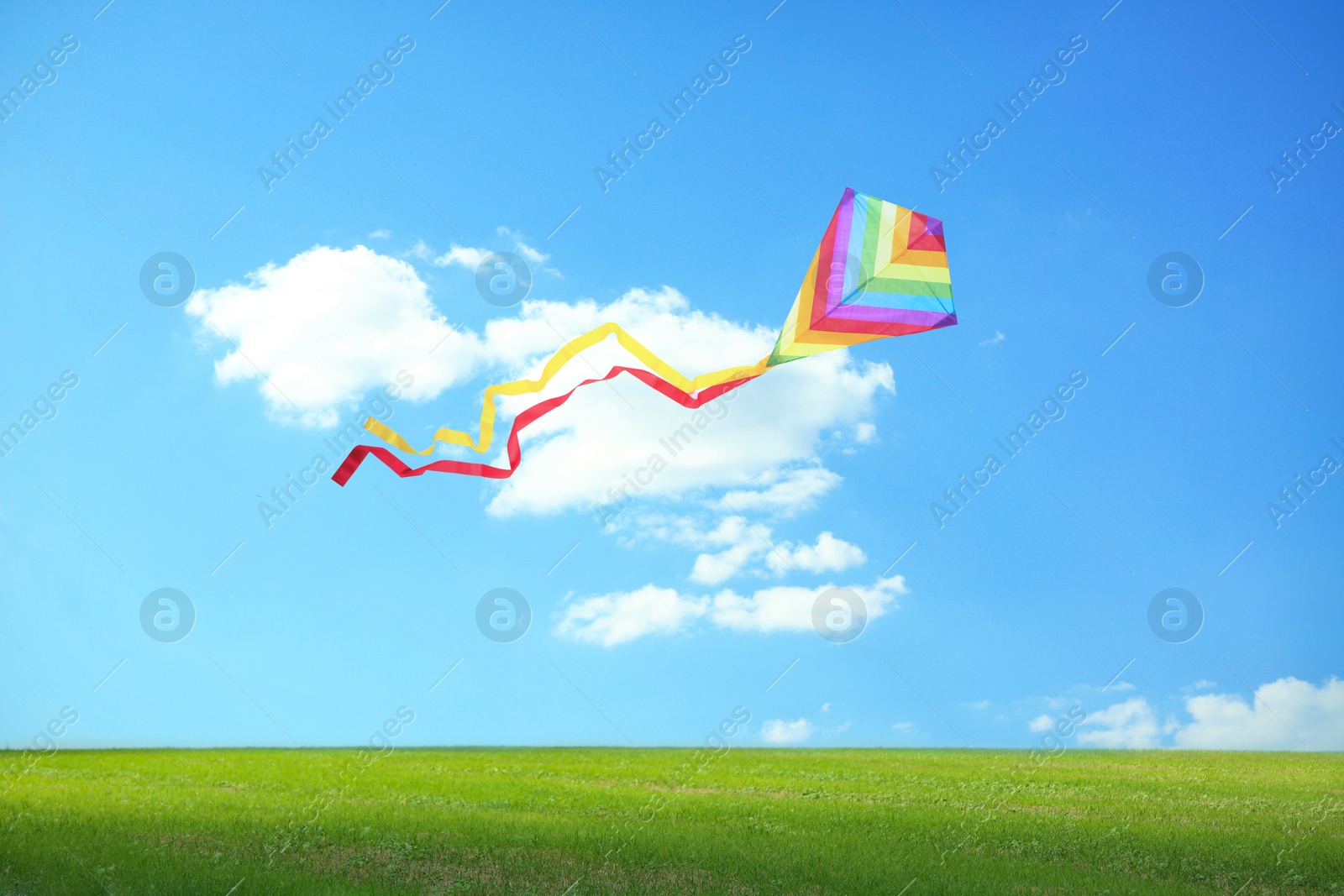 Image of Bright striped rainbow kite flying in blue sky over green grass on sunny day