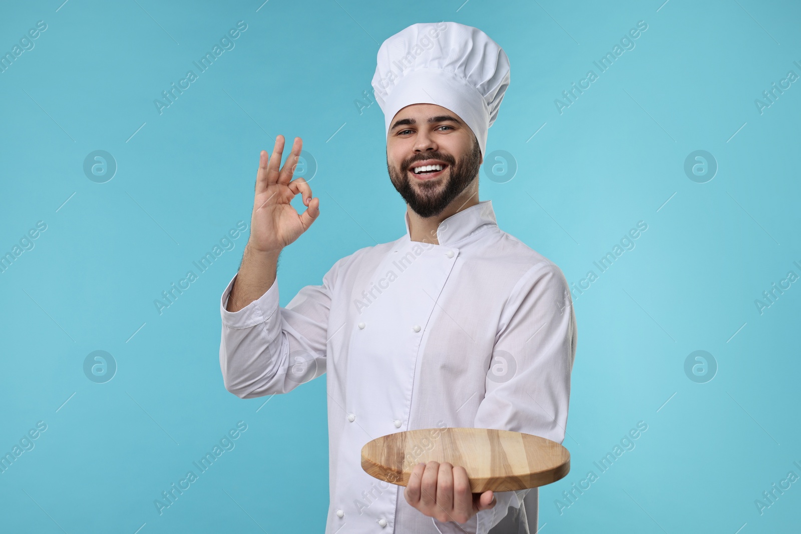 Photo of Happy young chef in uniform holding wooden board and showing ok gesture on light blue background