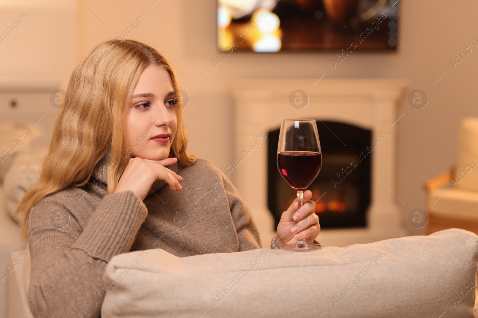 Photo of Beautiful young woman with glass of wine resting near fireplace at home