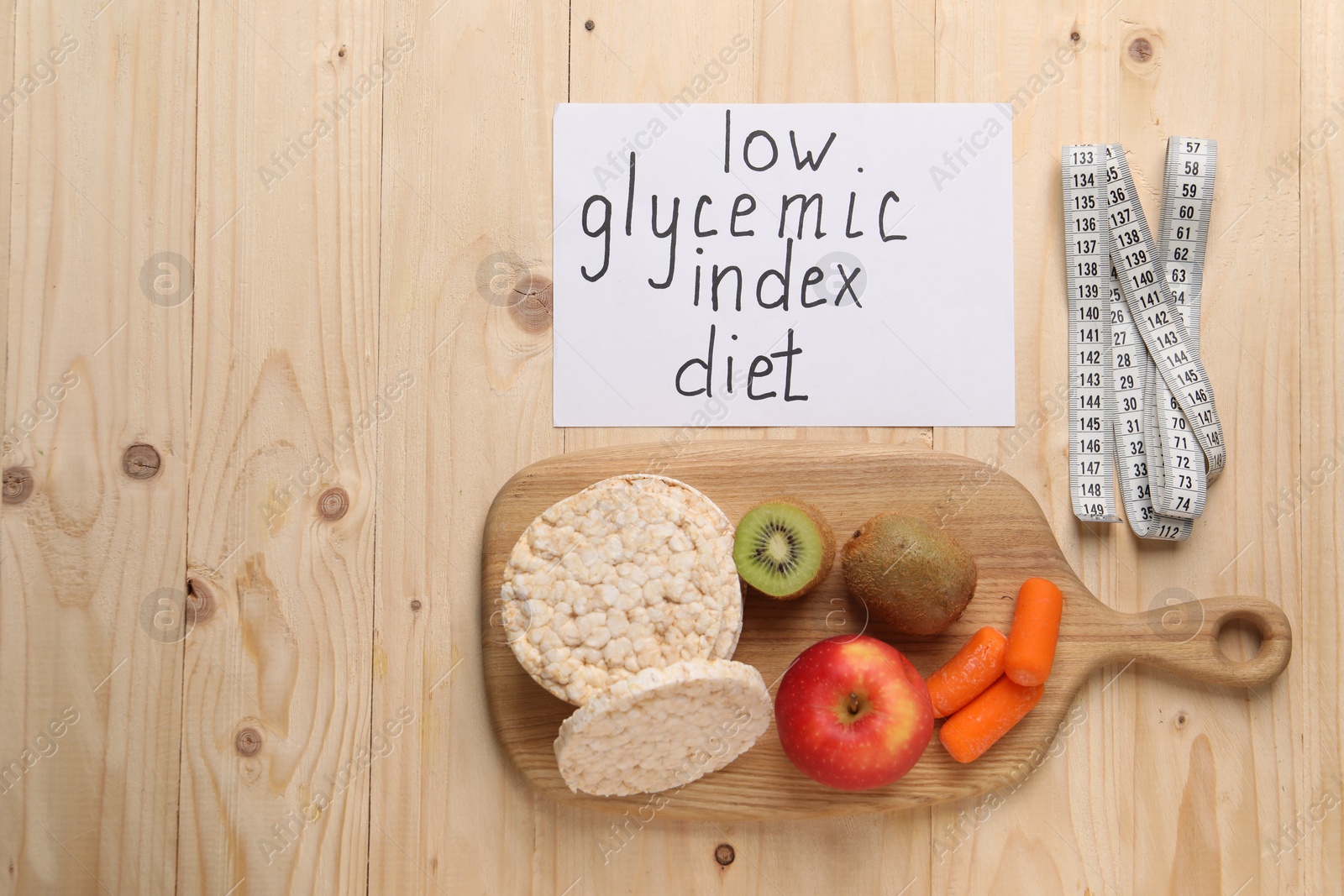 Photo of Paper with words Low Glycemic Index Diet, measuring tape and healthy products on wooden table, flat lay. Space for text