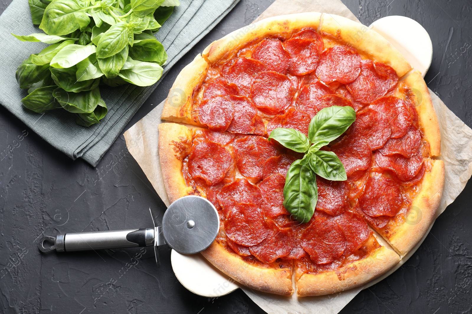 Photo of Tasty pepperoni pizza, cutter and basil on black textured table, flat lay