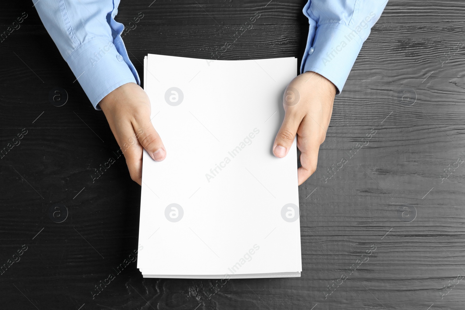 Photo of Man holding blank paper sheets for brochure at black wooden table, top view. Mock up