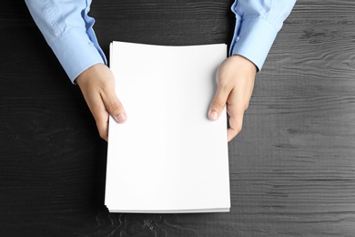 Man holding blank paper sheets for brochure at black wooden table, top view. Mock up