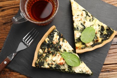 Pieces of delicious homemade spinach quiche, tea and fork on wooden table, flat lay