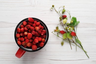Photo of Fresh wild strawberries in mug near stems with flowers and leaves on white wooden table, flat lay