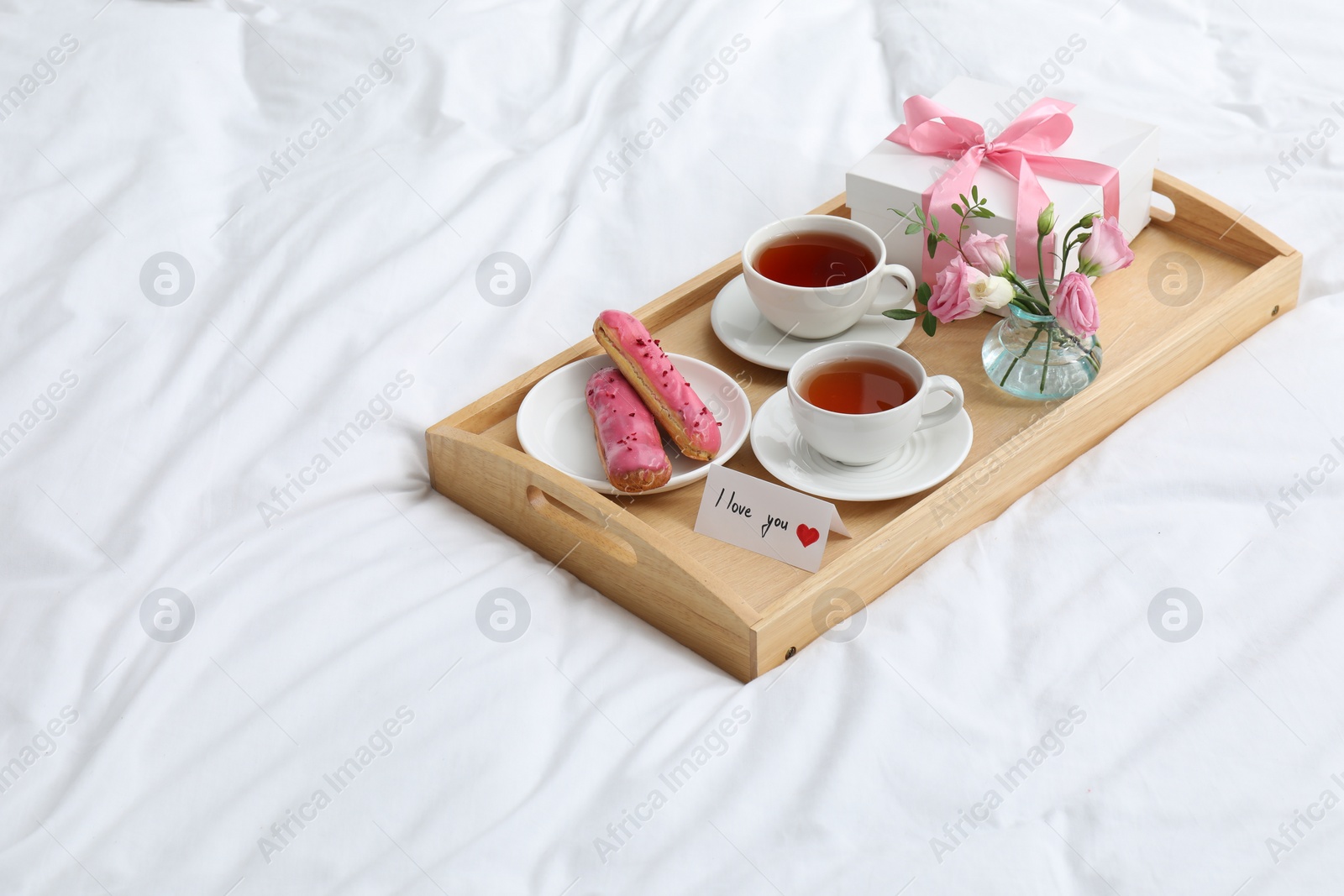 Photo of Tasty breakfast served in bed. Delicious eclairs, tea, gift box, flowers and card with phrase I Love You on tray, space for text