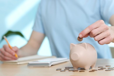 Financial savings. Man putting coin into piggy bank while writing down notes at wooden table, closeup