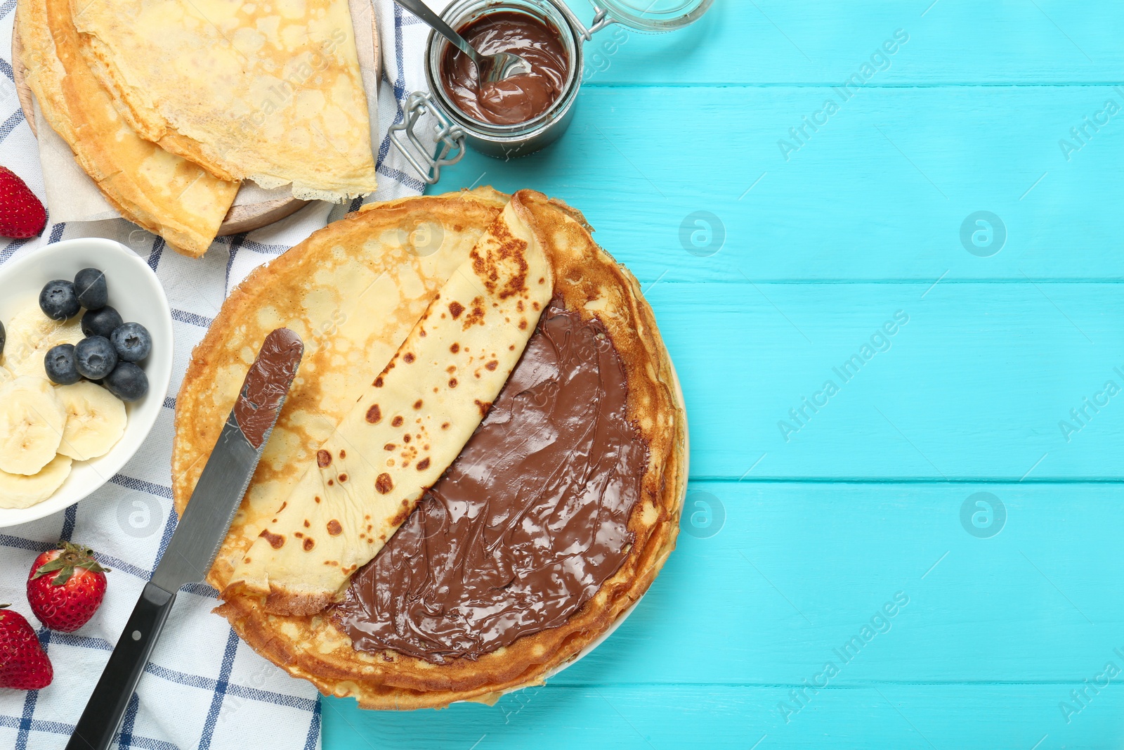 Photo of Tasty crepes with chocolate paste and berries served on turquoise wooden table, flat lay. Space for text