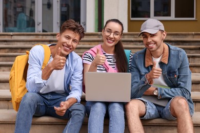 Happy young students with laptop showing thumbs up on steps outdoors