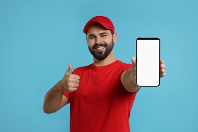 Image of Happy courier holding smartphone with empty screen and showing thumbs up on light blue background