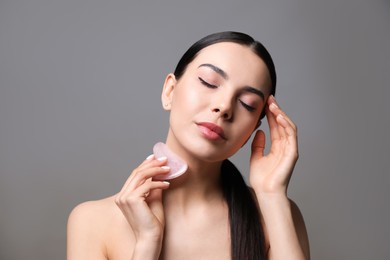 Photo of Beautiful young woman doing facial massage with gua sha tool on grey background