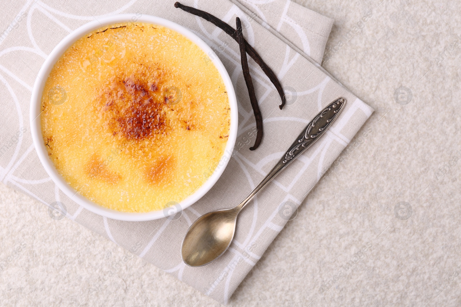 Photo of Delicious creme brulee in bowl, vanilla pods and spoon on light textured table, top view. Space for text