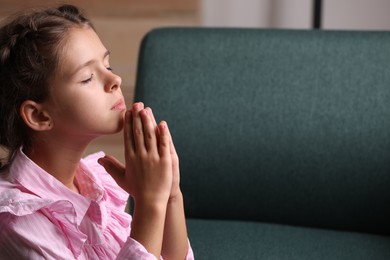 Photo of Cute little girl with hands clasped together praying at home. Space for text