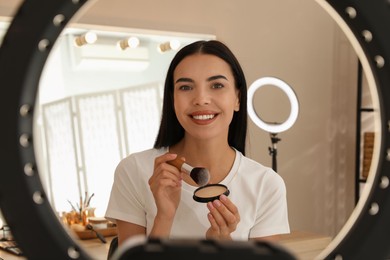 Photo of Beautiful young woman with face powder and brush indoors, view through ring lamp