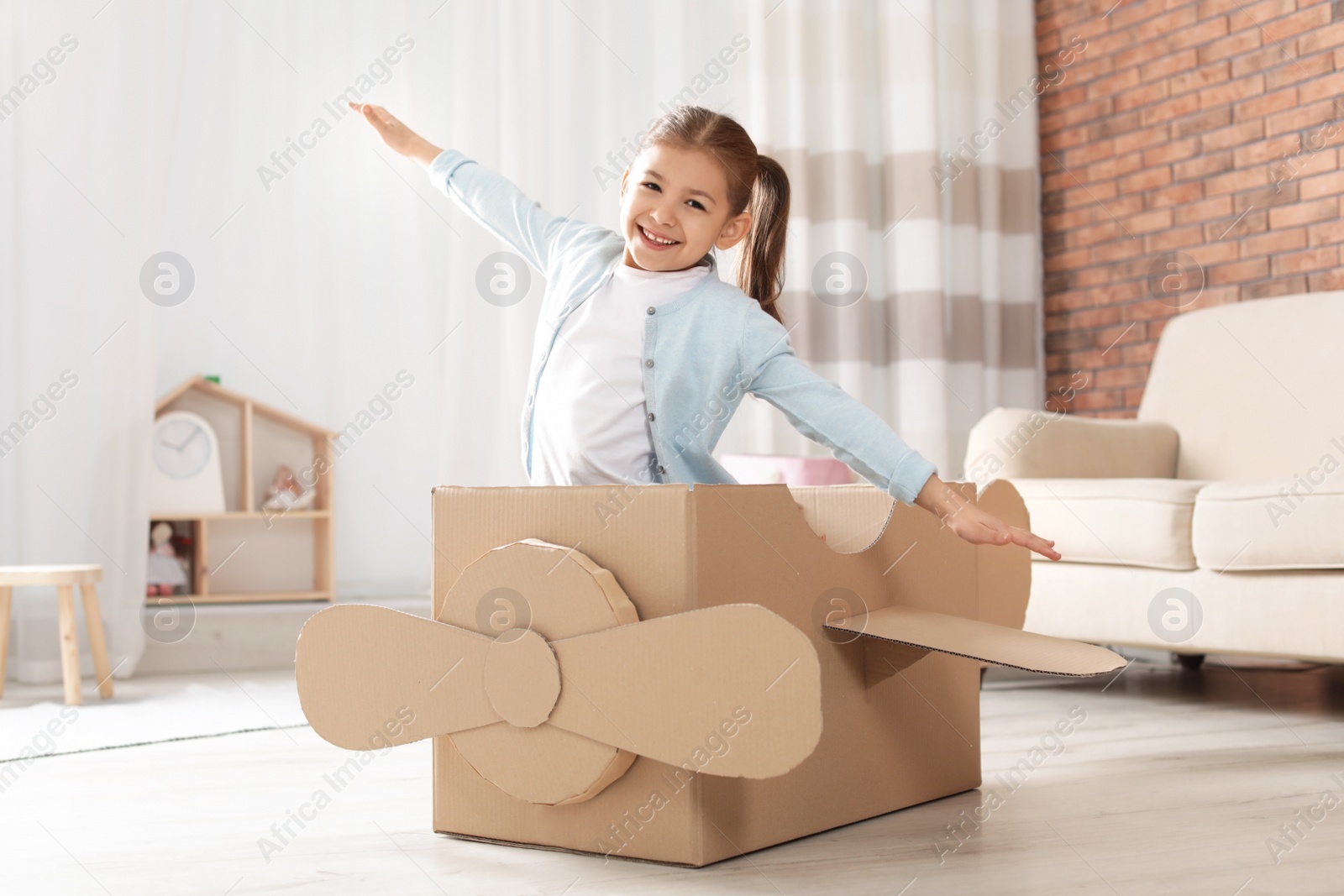 Photo of Cute little girl playing with cardboard airplane in living room