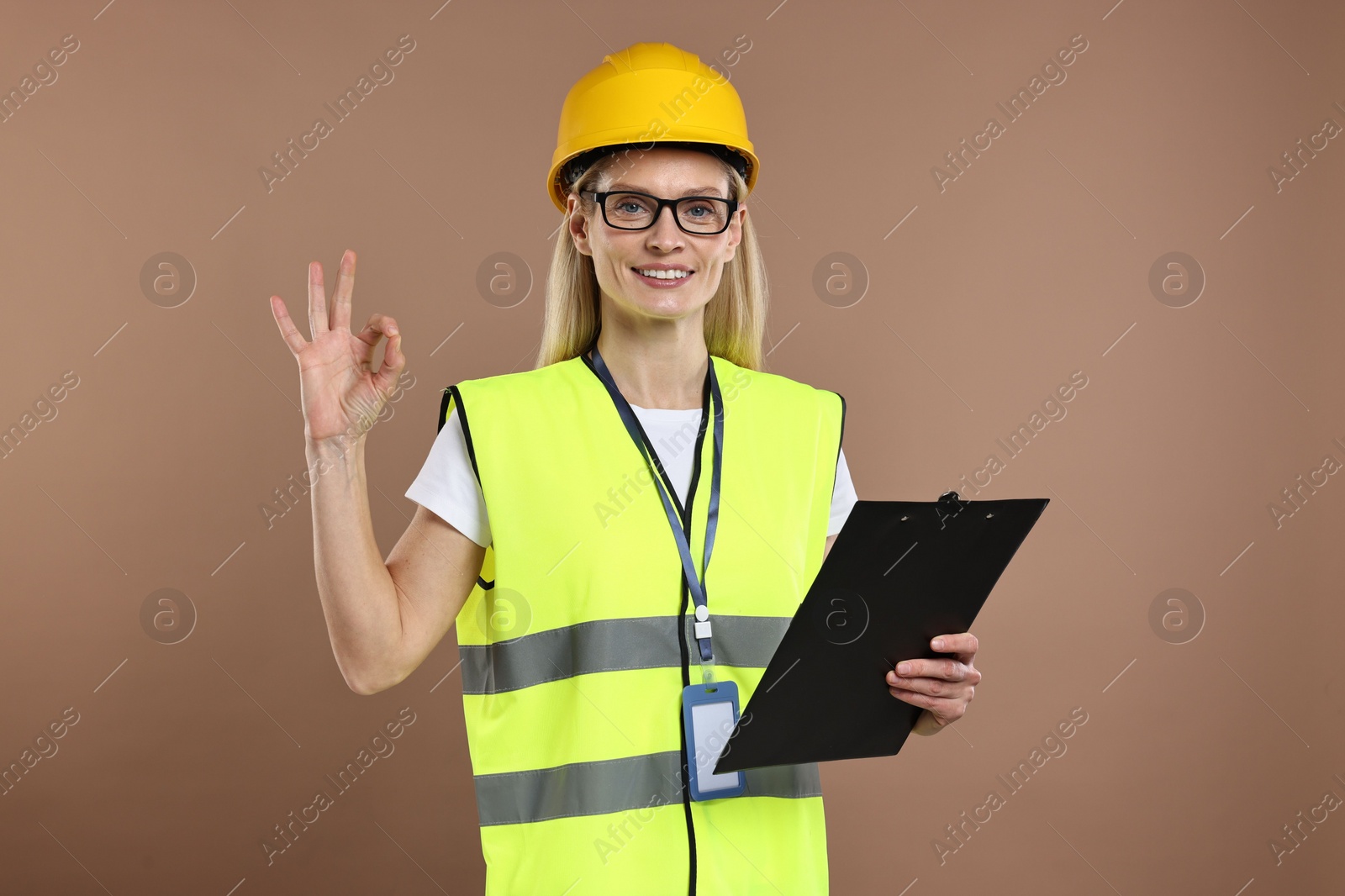 Photo of Engineer in hard hat holding clipboard and showing ok gesture on brown background