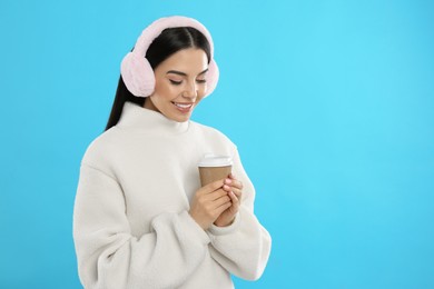 Beautiful young woman in earmuffs with cup of drink on light blue background. Space for text